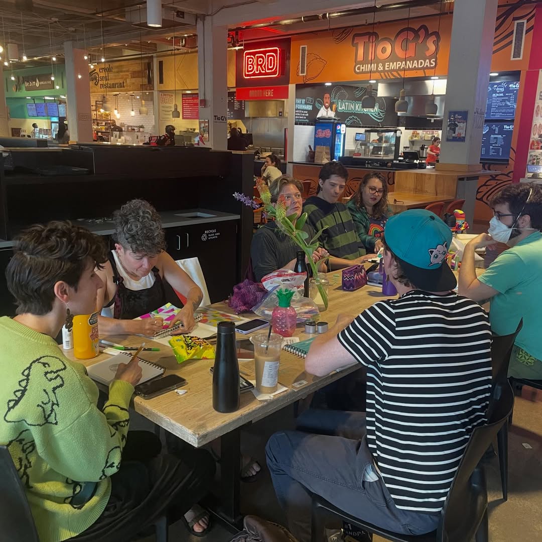 Drawing Club at a table in a food hall, sketchbooks and markers spread out among coffee cups.