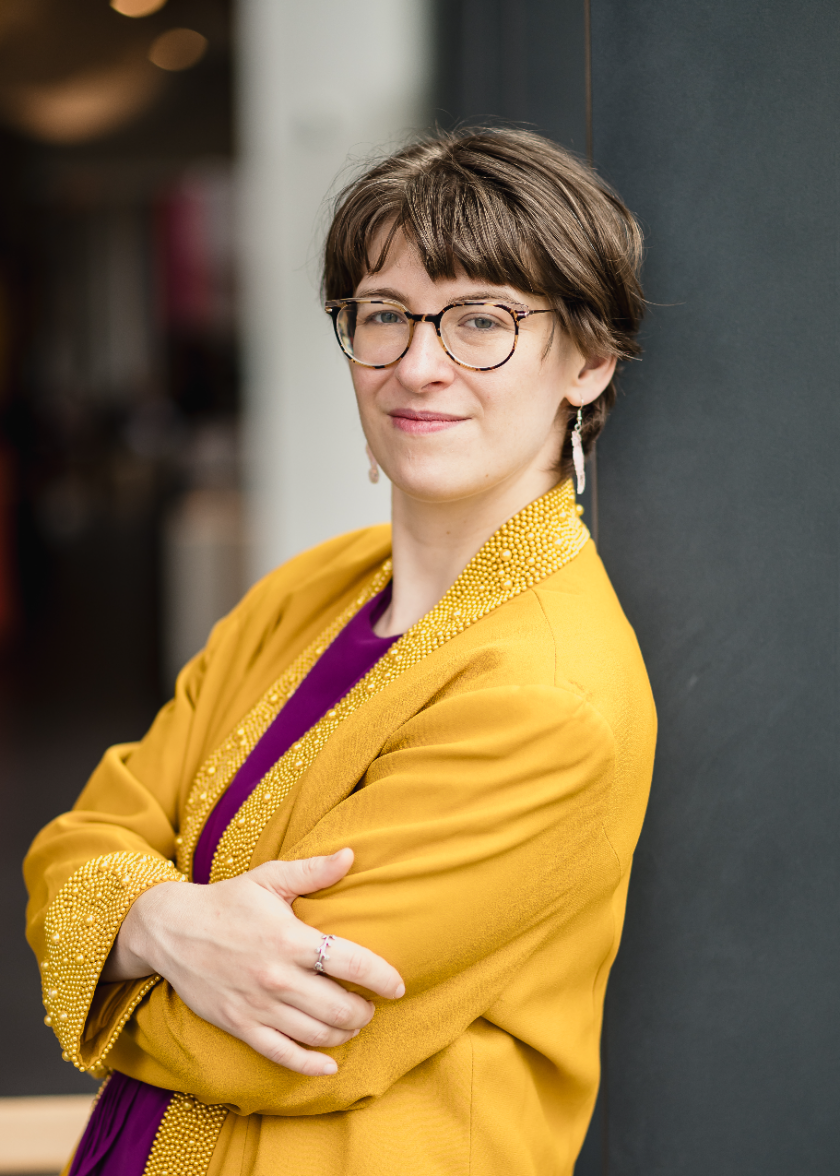 Portrait of Lehua Gray leaning against a dark grey wall, arms crossed, in tortoiseshell glasses and a mustard blazer with beaded lapels.