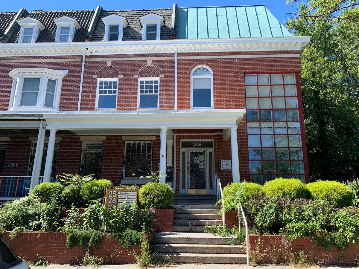The Lavender Collective building: a three-story brick rowhouse with a green copper roof, white porch columns, and a red-framed grid of windows on the right side.